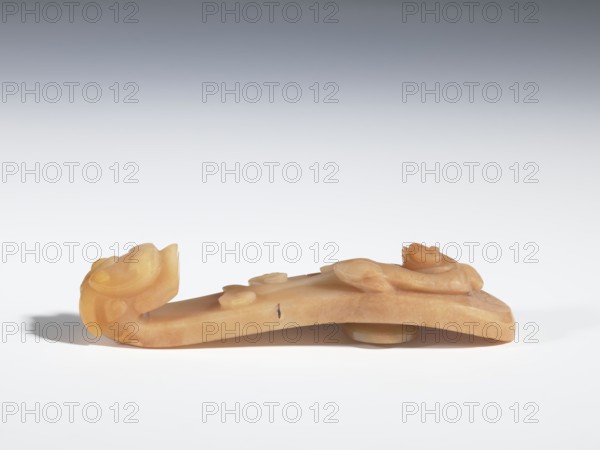 Belt clasp, Chinese culture, tigers zool., dragons iconographic, Qing 1644–1911. On the handle are engraved patterns in the shape of bats, a ruyi scepter head, and a leafy flower branch. Beige jadeite with darker spots. Carving, bas-relief, engraving; 18th–19th century, China. Belt buckle; height 2.4 cm, width 11.0 cm, depth 2.4 cm., A carved object in shades of pale tan and warm beige rests on a white surface against a gradient gray background, showing a horizontal, elongated form with rounded and curved carved elements at each end and a few raised rounded decorations along its top; the left end curls upward into a stylized, rounded shape while the right end also rises into a carved flourish, and a small circular protrusion is visible beneath the object near its right side, casting a soft shadow on the surface.