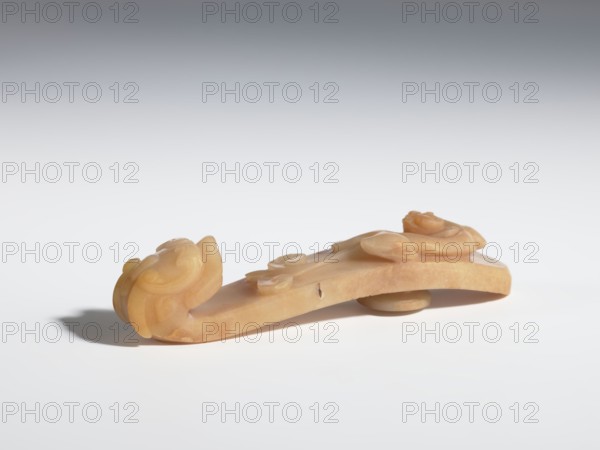 Belt clasp, purchase provenance; Chinese; tigers; dragons; Qing 1644–1911; On the handle are engraved patterns in the shape of bats, a ruyi scepter head, and a leafy flower branch. Beige jadeite with darker spots. Carving, bas-relief, engraving; 18th–19th century, China; belt buckle; height 2.4 cm, width 11.0 cm, depth 2.4 cm., A carved object made of a smooth, warm beige material rests on a white surface, showing a gently curved form with rounded, organic protrusions and flowing carved details; the object casts a soft shadow and has subtle variations in beige and light tan tones, with a gradual gray gradient background.