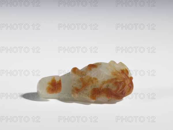Belt clasp, Chinese, dragons iconography, plant ornament, Qing 1644–1911, jade with chalcedony inclusions; bas-relief, engraving; fully carved head. 18th–19th century, China. Belt buckle; H 1.9 cm, W 6.2 cm, D 2.8 cm., A smooth carved object rests on a pale surface casting a soft shadow, the object showing a translucent whitish base with areas of warm amber-orange markings and patches, with rounded and flowing carved forms and raised textured details, set against a light gray to off-white gradient background.