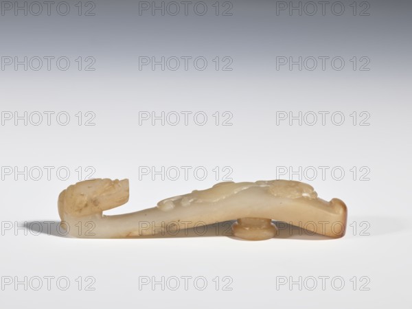 Belt clasp, Chinese, dragons iconography, Qing 1644–1911, bas-relief handle with a projection at the bottom — lizard-dragon chilong; head carved in full plastic. Jadeite with chalcedony inclusions. 17th–19th century, China. Belt buckle. Height 2.1 cm, width 10.6 cm, depth 1.8 cm., A carved object made of a pale, creamy stone with areas of light brown and amber tones lies horizontally on a flat white surface against a light gray gradient background; the object has a rounded raised base near one end and a curved elongated body with subtle carved bumps and shallow grooves, one end rising into a stylized head-like shape with carved facial features, and the opposite end curling slightly upward with a darker amber edge.