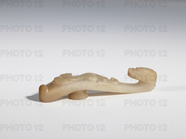 Belt clasp., Chinese; Qing dynasty 1644–1911. Bas-relief handle with a projection at the bottom — lizard-dragon chilong; head carved in full plastic. Jadeite with chalcedony inclusions. 17th–19th century. Belt buckle; height 2.1 cm, width 10.6 cm, depth 1.8 cm. Dragons iconography., A smooth, carved object in pale beige and light brown tones rests on a white surface with a soft shadow beneath; it has a long, curved form with a rounded, raised section near one end and a sculpted head-like shape at the other end, showing subtle carved contours and shallow relief details; the background is a gradient from white at the bottom to gray at the top.