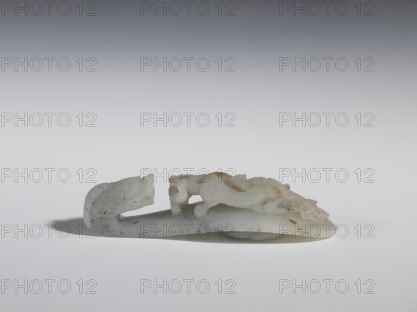 Belt clasp, purchase provenance. Chinese, Qing 1644–1911, 18th–19th century. Material: mossy chalcedony with openwork stem. Subject: lizard-dragon on a peony branch; underside knob shaped as a five-petaled flower rosette. Function: belt buckle. Dimensions: height 2.8 cm, width 11.0 cm, depth 3.8 cm., A pale carved object rests on a light surface against a gradient gray background, appearing to be made of an off-white material with areas of light brown and small dark speckles; the carving has smooth, curved forms suggesting a stylized animal-like figure with a raised head at one end and a sinuous body, casting a soft shadow beneath it.