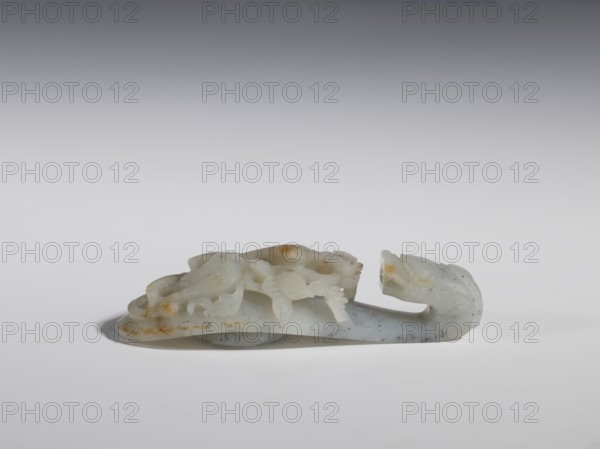 Belt clasp. Stem: lizard-dragon on a peony branch. Underside knob in the shape of a five-petaled flower rosette. Mossy chalcedony, openwork stem. Chinese, Qing dynasty 18th–19th century. Materials: chalcedony? Dimensions: height 2.8 cm; width 11.0 cm; depth 3.8 cm. Iconography: dragons, plant ornament., A pale carved object with smooth, curved forms rests on a light surface; the object shows shades of white, off-white, gray, and small areas of yellowish-brown discoloration, with a few darker gray speckles. The carving includes rounded, flowing shapes and a hooked end, with a group of overlapping, petal-like or leaf-like carved elements on the top. The background transitions from light gray near the surface to a darker gray toward the top of the image.
