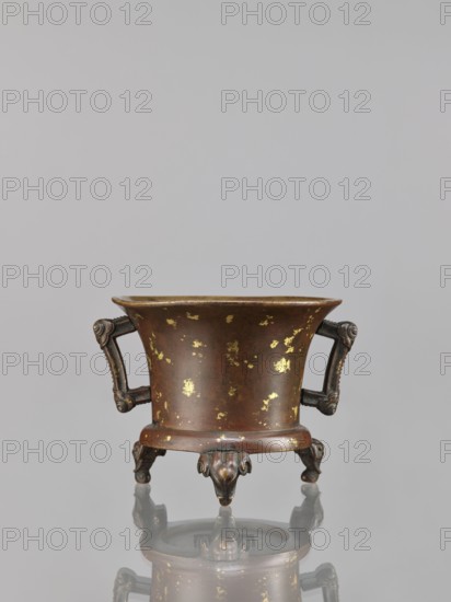 Censer, Chinese, Qing dynasty 1644–1911, 18th century. Bronze ritual/ceremonial vessel censer with elephant motifs; backdated signatures. Dimensions: height 9.7 cm, width 11.3 cm., A small metal vessel with a flared rim and a rounded body sits on ornate legs, showing a brown surface with scattered gold patches; it has a pair of decorative handles with curled ends and sculpted detailing, and the three legs terminate in stylized animal-like feet, all reflected faintly on a smooth gray surface beneath; the background is a uniform light gray.