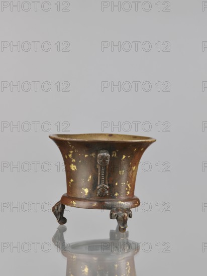 Censer, Chinese, Qing dynasty 1644–1911, 18th century. Ceremonial/ritual vessel; backdated signatures. Material/subject: vessel censer with elephants. Dimensions: height 9.7 cm, width 11.3 cm., A small metal vessel with a flared rim stands on a short rounded base supported by three sculpted feet shaped like stylized animal heads; the surface shows worn patches of gold over a brownish-bronze patina with areas of darker tarnish, and the interior appears lighter with a muted golden tone, all reflected faintly on the glossy surface beneath.