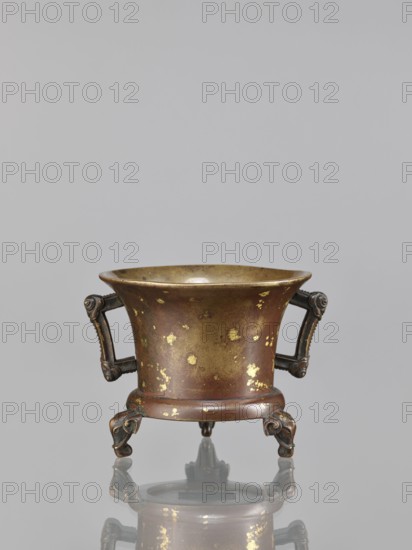 Censer, Chinese, Qing 1644–1911, 18th century, China; ceremonial/ritual bronze vessel with backdated signatures, decorated with elephants; height 9.7 cm, width 11.3 cm., A small metal vessel with a flared rim and a rounded body sits on a reflective surface; its surface shows patches of worn gilding giving gold flecks over a brown and bronze base color. The vessel has a pair of ornate handles on opposite sides, each handle dark bronze with decorative scrollwork, and it stands on three short, sculpted legs that curve outward and have darker bronze tones. The interior of the vessel appears smoother and more golden than the exterior, and the reflection beneath echoes the shape and colors of the metal object. The background is a smooth, even light gray.