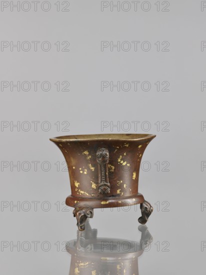 Censer, Chinese art, Chinese culture, Qing 1644–1911, ceremonial vessels, ritual vessels, backdated signatures, elephants, 18th century, China, vessel, censer, height 9.7 cm, width 11.3 cm, A small decorative metal vessel with a flared rim sits on a reflective surface, showing a mirrored reflection below; the vessel is brown with patches of gold leaf or gilding scattered across its surface and has darker brown decorative vertical mounts and ornate feet with curled, sculpted forms, while the interior rim appears slightly lighter and the background is a smooth pale gray.