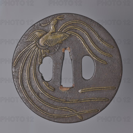 Crossguard tsuba, Japanese, Edo period 17th–18th century. Round tsuba maru gata with a slightly rounded edge kaku mimi ko-niku, copper sekigane. Decorated in low relief with brass inlay depicting two phoenixes hôô in flight on one side and one phoenix on the other, with decoratively spread long tails. Not signed. Diameter 8.5 cm; thickness 0.5 cm., A round metal object with a dark brown surface and golden inlaid lines forming the shape of a bird with flowing tail feathers, showing a bird with outstretched wings near the top and long curved feathers that sweep across the face; there are two rounded cutouts and one narrow central cutout interrupting the surface, and the metal shows areas of wear with hints of reddish-brown and copper tones where the surface is exposed.