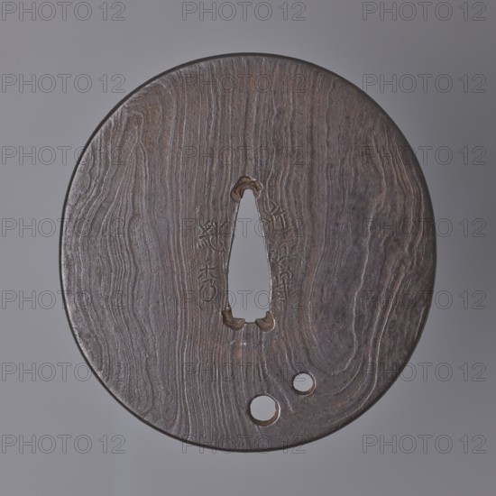 Crossguard tsuba, Tsuguhide. Oval tsuba tatemaru gata with a kaku mimi ko-niku type edge. Flat, forged from many layers, with two round holes udenuki ana, surface shaped like a tree trunk grain pattern mokume ji decoration. Signed. Note: the word kami lit. prince, lord is one of the titles used by tsuba masters and often included in signatures. Early 19th century, Japan, Ômi Eshu. Tsuba. Height 8.2 cm, width 7.7 cm, depth 0.4 cm., A round, flat object with a textured wood-like surface in shades of brown and dark brown sits against a neutral gray background; its center has a vertical, teardrop-shaped opening with rough, slightly darker edges and a few small carved notches nearby, and toward the lower edge there are a few circular holes; the surface shows flowing grain patterns and subtle variations of brown, dark brown, and hints of gray.
