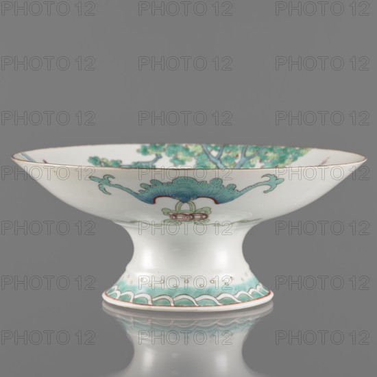 Platter, porcelain with white transparent glaze, decoration painted with overglaze enamels and iron red, gilded edge. Qing dynasty, Tongzhi period 1862–1874. Chinese; colored porcelain depicting go game, figural scenes of women, scholars, and musical instruments. Porcelain vessel, height 8.5 cm, diameter 21 cm., A shallow pedestal bowl made of white porcelain with a flared rim and a rounded foot, decorated with painted organic motifs in shades of green and turquoise with accents of red and pale gold; the interior rim shows a band of green foliage, while the exterior displays stylized floral and wave-like patterns in green and turquoise with small red highlights and delicate pale gold edging; the pedestal foot also features a decorative green and turquoise border with tiny gold dots, and the bowl sits on a reflective gray surface against a solid gray background.