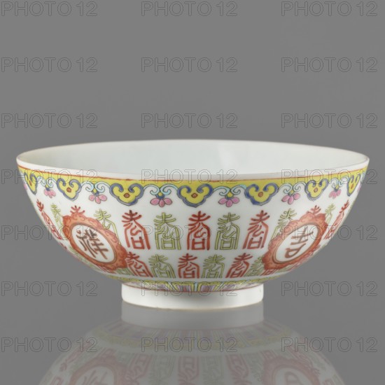Bowl. Porcelain body with white glaze; decoration painted with overglaze enamel paints and iron red. Colored porcelain, lambrequin pattern, longevity shou iconography. Jiaqing period early 19th century, 1796–1820, Qing dynasty 1644–1911, China. Bowl on a ring foot. Height 5.8 cm; diameter 14.15 cm., vessel., A shallow porcelain bowl with a rounded rim and a short foot, shown against a neutral gray background, decorated around the exterior with a repeating ornate pattern in red, green, yellow, blue, pink, and a touch of orange, featuring stylized vertical motifs and framed circular medallions, a yellow and blue cloud-like border near the rim, and a pale off-white interior and base; the bowl sits on a reflective surface that mirrors its colors and pattern.