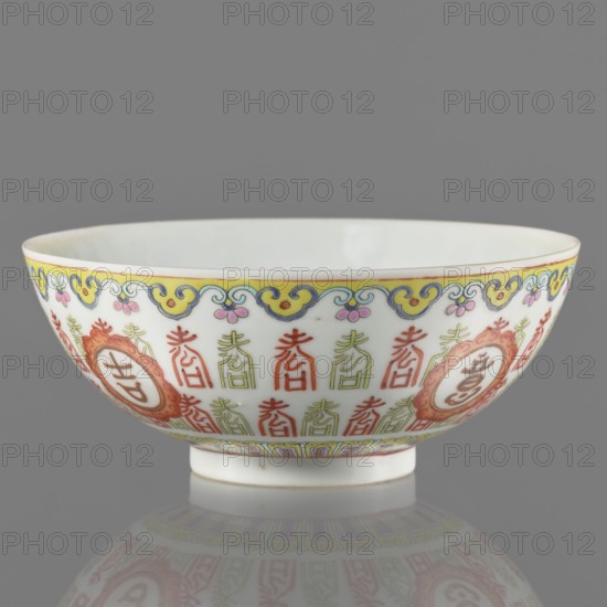 Bowl. Porcelain body with white glaze; decoration painted with overglaze enamel paints and iron red, featuring longevity shou iconography and lambrequin pattern in Jiaqing style Qing dynasty. Early 19th century Jiaqing period, 1796–1820. China. Vessel on a ring foot. Height 5.8 cm; diameter 14.15 cm., Colored porcelain; Chinese art; Qing 1644–1911., A porcelain bowl shown in profile with a glossy white interior and exterior, decorated around the rim with a yellow band and a scrolling cloudlike border in blue and yellow with small pink floral motifs; the exterior body features repeating vertical ornamental motifs in red and pale green, framed by circular cartouches in orange-red with decorative surrounds, and a narrow decorative band in soft yellow and green near the foot; the bowl sits on a short white foot and is reflected faintly on the smooth gray surface beneath; visible colors include white, yellow, blue, pink, red, orange, green, and gray.