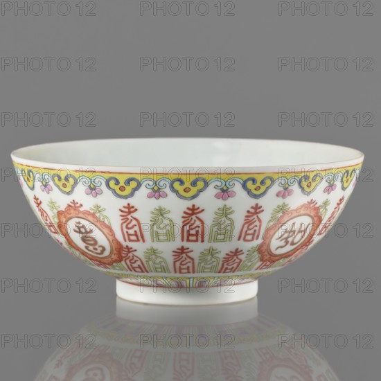 Bowl. Porcelain body with white glaze; decoration painted with overglaze enamel paints and iron red. Colored porcelain with longevity shou iconography and lambrequin pattern in Jiaqing style, Qing dynasty, early 19th century Jiaqing period, 1796–1820. Bowl on a ring foot. Height 5.8 cm; diameter 14.15 cm., ceramics; Chinese., A shallow porcelain bowl with a white interior and exterior, decorated around its rim and sides with a continuous painted border in yellow, blue, pink, green, and red; the upper border features a repeating cloud-like motif in yellow and blue with small pink floral accents, while the main band below shows a repeated vertical ornamental pattern in green and red interspersed with round medallions framed in red and orange; the foot rim has a narrow painted band in pale green and red with geometric detailing; the background behind the bowl is gray and the bowl casts a soft reflection on a glossy surface beneath it.