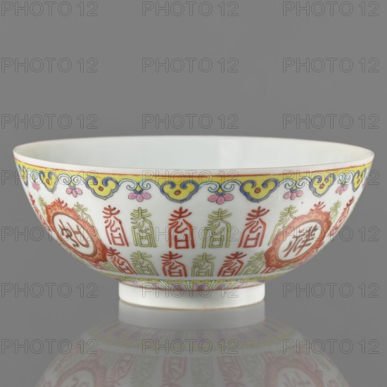 Bowl, porcelain body with white glaze; decoration painted with overglaze enamel paints and iron red featuring longevity shou iconography and lambrequin pattern in Jiaqing early 19th century, 1796–1820 style, Qing dynasty 1644–1911; bowl on a ring foot; colored porcelain; China; vessel; height 5.8 cm, diameter 14.15 cm., A porcelain bowl sits on a reflective surface against a gray background, showing a white interior and exterior decorated with a decorative border near the rim in yellow, blue, pink, and a thin red line; the exterior features repeating vertical motifs in red and green and circular medallions in red with ornamental frames, plus a lower band of geometric pattern in yellow, blue, pink, and green; the bowl has a small white foot and the reflection beneath shows muted colors from the decoration.