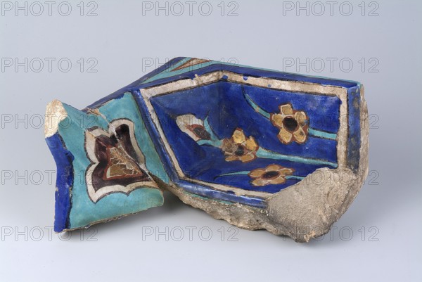 Fragment of a mukarnas, Islamic art, Persian culture, Timurid dynasty 1370–1526, Timur 1336–1405, architecture — mosques, fragments, architectural element, palmettes and floral ornament, glued, surface abrasions, end of the 14th century, Turkestan Kazakhstan, architectural element; height 21.4 cm, width 20.5 cm, depth 10.3 cm., A broken ceramic fragment with a glossy surface showing a deep blue interior decorated with floral motifs in gold, brown, turquoise, and black, bordered by a lighter blue edge and an off-white rim; the outer surface displays turquoise and cobalt blue areas with a stylized heart-shaped design in brown, cream, and black, and the piece rests on rough, unglazed beige-gray clay with some worn and chipped sections.