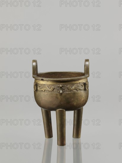 Ding incense burner, leiwen ornament, Chinese art, Ding vessel, Chinese culture, ceremonial/ritual vessel, birds decorative motif, meander ornament, Qing 1644–1911, 19th century, China, vessel, height 19.2 cm, diameter 13.6 cm, A metal tripod vessel with a rounded body and a pair of upward-curving loop handles sits against a plain light gray background; the vessel shows a metallic bronze and dark brown patina with areas of greenish tarnish and subtle reddish-brown spots, a decorative band with raised curvilinear motifs encircles the upper body, and three cylindrical legs extend downward, each reflecting slightly on the smooth surface beneath.