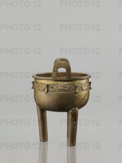 Ding incense burner, leiwen ornament, Chinese art; Ding vessel; Chinese; ceremonial/ritual vessel with bird decorative motif and meander ornament; Qing dynasty 1644–1911, 19th century, China; vessel; height 19.2 cm, diameter 13.6 cm, A small tripod vessel made of a dull gold-colored metal with a rounded body and a flat rim, supported by three cylindrical legs, one of which shows a patch of reddish-brown material; the upper band of the body features a decorative raised pattern and small tab-like projections, and a large arch-shaped handle rises from the rim; the background is a smooth light gray and the surface beneath the vessel reflects it faintly.