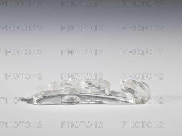 Belt clasp, Chinese; Qing 1644–1911; 17th–19th century. Material: rock crystal with inclusions. Subject/iconography: lizard-dragon chilong. Description: full-plastic carved head; shank with deep relief, openwork, and engraving. Function: belt buckle for men's clothing. Dimensions: height 2.0 cm, width 9.8 cm, depth 2.0 cm., A translucent carved object rests on a smooth surface, showing clear and frosted areas with glossy highlights; its elongated shape curves upward at one end into a rounded, leaflike form with carved grooves, while the opposite side features low-relief rounded motifs and gentle undulations; the piece casts a soft shadow and the visible colors are clear, white, gray, and subtle silvery tones.