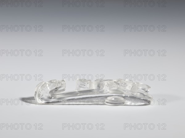 Belt clasp, purchase provenance; Chinese; dragons iconography; Qing 1644–1911; clothing; belts; men's clothing; costumes; ornaments; men's ornaments; lizards zool.; buckles. Shaft: lizard-dragon chilong. Rock crystal with inclusions; full-plastic carved head; shank: deep relief, openwork, engraving. 17th–19th century, China. Belt buckle. Height 2.0 cm; width 9.8 cm; depth 2.0 cm., A transparent glass or clear plastic decorative object rests on a smooth light gray surface against a gradient gray background, showing a curved elongated base with an upward-arching sculpted end and a series of raised, rounded forms along its top; the object casts soft pale gray shadows and displays reflections and highlights in clear, colorless tones.