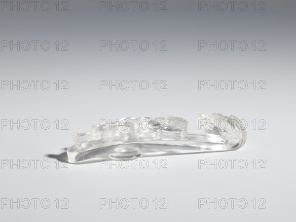 Belt clasp, Chinese; Qing 1644–1911. Rock crystal with inclusions; full-plastic carved head; shank: deep relief, openwork, engraving. Shaft: lizard-dragon chilong. Dragons iconography; lizards zool.. Function: belt buckle for men's clothing/ornaments, costumes. 17th–19th century. Dimensions: height 2.0 cm, width 9.8 cm, depth 2.0 cm., A clear, transparent carved object rests on a smooth, light gray surface with a gradient gray background; the object has curved, elongated forms with flowing, leaf-like and rounded ornamental shapes, showing glossy reflections and soft shadows beneath it, and the visible colors are clear/transparent, gray, and white.