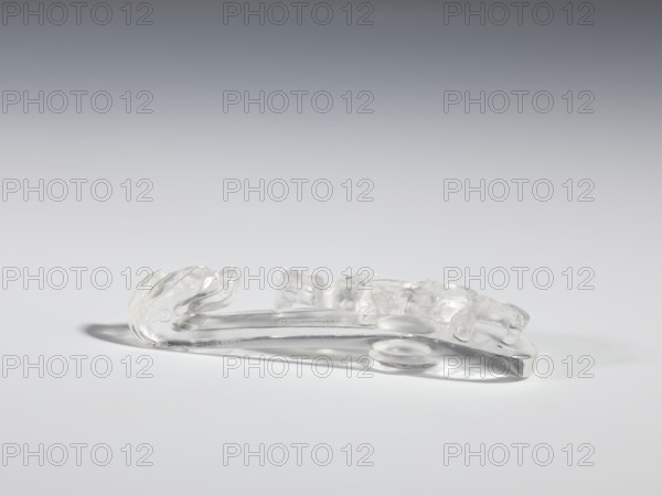 Belt clasp, Chinese; Qing 1644–1911; 17th–19th century. Materials and form: shaft: lizard-dragon chilong in rock crystal with inclusions; full-plastic carved head; shank with deep relief, openwork, and engraving. Function: belt buckle for men's clothing/ornaments. Dimensions: height 2.0 cm, width 9.8 cm, depth 2.0 cm. Iconography: dragons, lizards., A clear, translucent object rests on a smooth light-gray surface against a gradient gray background, showing a curved elongated base with a sculpted, sculptural end that resembles flowing shapes; the object casts a soft shadow and displays highlights and reflections in shades of white, clear, and pale gray.