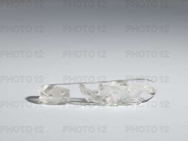 Belt clasp, Chinese, Qing 1644–1911. Shaft: lizard-dragon chilong. Rock crystal with inclusions; full-plastic carved head; shank: deep relief, openwork, engraving. 17th–19th century. Belt buckle. Height 2.0 cm; width 9.8 cm; depth 2.0 cm. Subjects/iconography: dragons, lizards; function: men's clothing ornament belt buckle., A clear translucent carved object rests on a smooth surface against a background that fades from light gray to darker gray; the object is colorless and glass-like with glossy highlights and soft shadows, featuring flowing, curved sculpted forms and rounded ends, one end showing a stylized head-like shape and the body displaying sinuous, wave-like carvings and subtle textured grooves, with reflections and refractions creating areas of white sheen and pale gray tones.