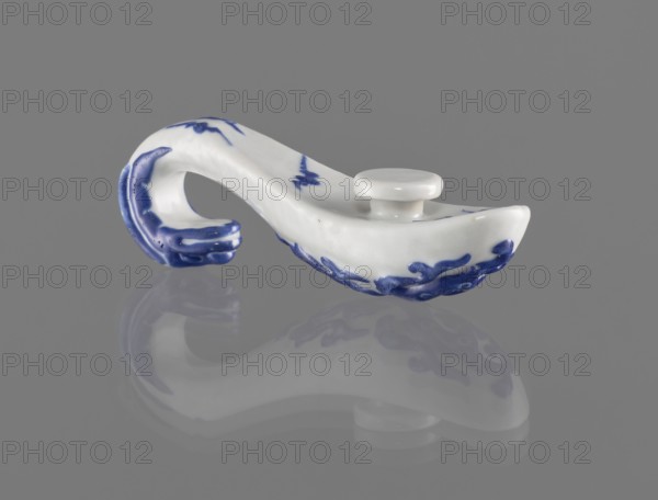 Belt clasp, Chinese; 19th century. Porcelain belt buckle decorated with navy blue overglaze enamel paint. Shaft: stylized lizard-dragon; bats on the underside. Materials: colored porcelain, ceramics. Dimensions: height 3.1 cm, width 9.3 cm, depth 2.65 cm. Icons: dragons, bats., A ceramic object with a curved handle and a shallow bowl-like end sits on a smooth gray surface, showing a soft reflection beneath it; the piece is primarily white with decorative blue painted accents along the handle and rim, and a small white circular knob on top of the bowl area; the background is uniform gray.