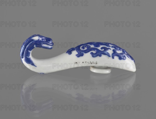 Belt clasp., Chinese, 19th century. Shaft: stylized lizard-dragon, bats on the underside. Navy blue overglaze enamel paint on colored porcelain. Function: belt buckle. Dimensions: height 3.1 cm, width 9.3 cm, depth 2.65 cm., A small curved ceramic object shaped like an animal with a curled neck and head, resting on a flat reflective surface, is shown against a plain gray background; the object is primarily white with areas of deep blue forming a stylized pattern on its back and blue detailing on the head and neck, and its reflection in the surface mirrors the white and blue colors.