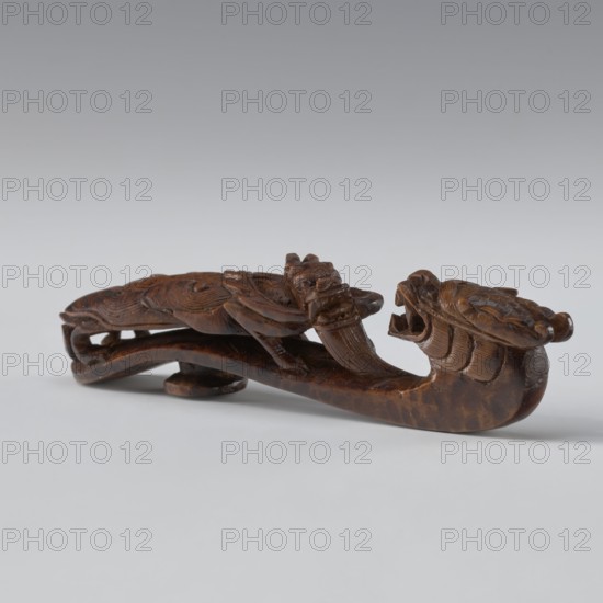 Belt clasp, maker, Chinese; 19th century Qing, 1644–1911. Materials and technique: full-sculpture carved head with openwork shank. Subject/iconography: two fighting lizard-dragons on the shaft. Function: men's clothing ornament belt clasp. Dimensions: height 3.0 cm; width 12.8 cm; depth 2.8 cm., A small carved wooden object in brown tones sits on a light gray surface against a pale gray background; the carving features a stylized animal head with an open mouth at one end and a curled tail or body with carved feather or scale-like textures along its length, showing a few raised ridges and carved details, with visible wood grain and darker brown variations across the surface.