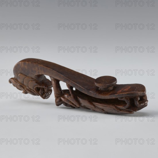 Belt clasp, Chinese, Qing 1644–1911, 19th century. Material/technique: full-sculpture carved head; openwork shank. Subject/iconography: two fighting lizard-dragons. Function: clothing ornament for men's belts. Dimensions: height 3.0 cm, width 12.8 cm, depth 2.8 cm., A carved wooden object in shades of brown rests on a pale gray surface against a lighter gray background, showing a curved elongated form with a rounded raised knob on top and intricate carved details along its underside and sides, including a series of carved ridges and ties that create textured patterns and hollowed areas, with the wood surface displaying darker brown areas and lighter brown highlights.