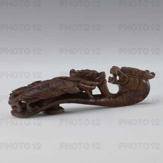 Belt clasp, Chinese; dragons iconography; Qing 1644–1911; 19th century; men's clothing ornament — belts. Shaft: two fighting lizard-dragons. Full-sculpture carved head; openwork shank. Materials/technique: carved. Dimensions: height 3.0 cm, width 12.8 cm, depth 2.8 cm., A carved wooden object in dark brown tones shaped like a stylized creature with an elongated curved body, a pronounced open-mouthed head with visible teeth, and intricately carved flowing details along its back and sides, resting on a light neutral surface against a soft gradient background of pale gray to muted bluish-gray.