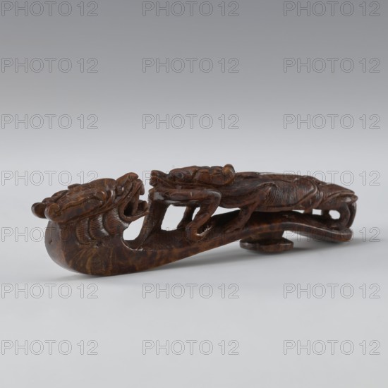 Belt clasp, Chinese; Qing dynasty 1644–1911, 19th century. Materials/technique: full-sculpture carved head, openwork shank. Subject/iconography: two fighting lizard-dragons. Function: men's clothing ornament belt. Dimensions: height 3.0 cm, width 12.8 cm, depth 2.8 cm., A carved wooden object depicting a stylized animal form with a long curved body and open mouth, shown resting on a flat surface; the carving has detailed textured areas and interlocking limbs or appendages, with visible grain and carved grooves. The colors visible are brown in various tones on the carving and shades of gray and white in the background and surface.