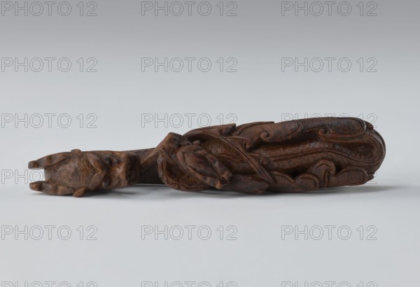 Belt clasp, Chinese, Chinese art; dragons iconography; Qing 1644–1911; men's clothing ornament belt; shaft: two fighting lizard-dragons; full-sculpture carved head; openwork shank; 19th century, China; height 3.0 cm, width 12.8 cm, depth 2.8 cm., A carved wooden object rests horizontally on a light gray surface against a pale gray background, showing a dark brown color with visible carved patterns and textures; the carving features a stylized animal head at one end with textured scales and small protruding ears, a twisted or knotted central section with interlaced carved elements, and flowing, curved motifs along the body that resemble leaves or waves, all rendered in varying shades of brown with subtle highlights and darker recesses.