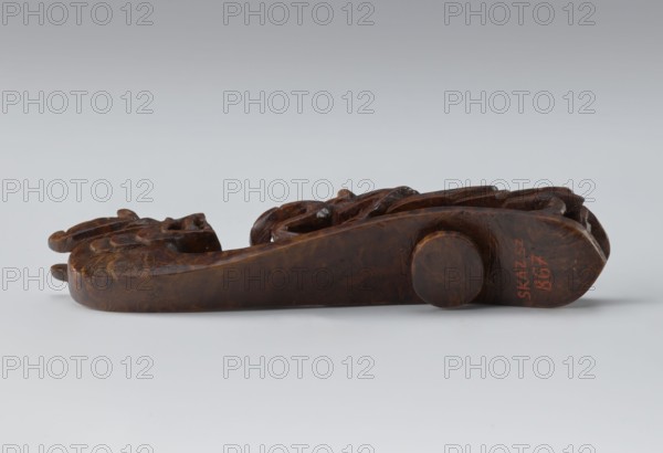 Belt clasp, Chinese; Qing 1644–1911, 19th century; shaft: two fighting lizard-dragons; full-sculpture carved head; openwork shank; materials: carved unspecified; function: clothing/belt ornament for men's costume; height 3.0 cm, width 12.8 cm, depth 2.8 cm; dragons iconography., A carved wooden object rests horizontally on a pale gray surface against a light gray gradient background; the object is dark brown with varied wood grain and carved details including rounded and raised forms, smooth flat sides, and textured carved sections, and there is a small circular wooden peg on one side, with visible shades of brown and touches of darker brown in the carved grooves and a faint reddish-brown area.