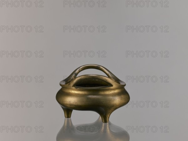 Li-type incense burner, Li-type vessel, Qing dynasty 1644–1911, 18th century, China. Bronze vessel in Li form used as an incense burner, decorated with fantastical beasts; backdated signatures; height 7.5 cm, width 12.1 cm., A small metallic vessel with a rounded body and a warm brass tone sits on a reflective surface, supported by three short tapered legs and topped by a smooth curved double-handle arch; the vessel shows subtle surface variations and patina in shades of brass, gold, and darker brown, while the background is a soft neutral gray.