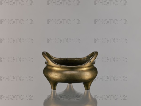 Li-type incense burner, Chinese, ceremonial Li-type vessel, Qing dynasty 1644–1911, 18th century China, backdated signatures, fantastical beasts, ritual vessel, incense burner, height 7.5 cm, width 12.1 cm, A small metallic vessel with a warm brass color sits centered on a reflective surface, showing a soft reflection below. The vessel has a rounded body with a slightly bulging midsection, a wide open top with two upward-curving handles on opposite sides, and three short tapered legs. The surface of the metal shows subtle darker patina spots and small areas of wear. The background is a smooth, neutral gray with gentle gradient shading. The visible colors are brass, dark brownish patina, gray, and muted reflections of those tones.