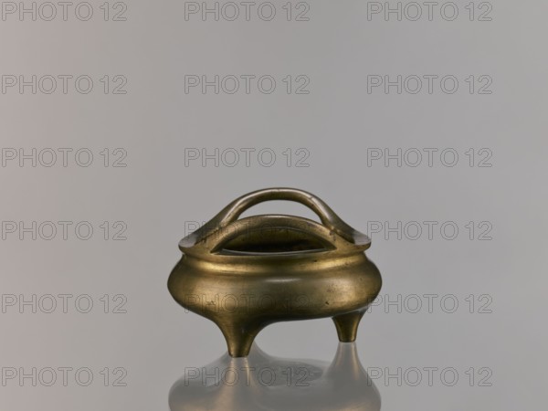 Li-type incense burner, Chinese art, Chinese culture, ceremonial vessel, Li-type vessel, Qing 1644–1911, fantastical beasts, ritual vessel, Qing style, backdated signatures, 18th century, China, vessel, incense burner, height 7.5 cm, width 12.1 cm, A small rounded metal vessel with a warm brass color sits on a reflective surface, showing a soft reflection below; the vessel has a bulbous body tapering into three short rounded legs and features a double-arched handle that rises above an open central rim, with the metal surface displaying subtle darker spots and a slightly aged patina against a plain light gray background.