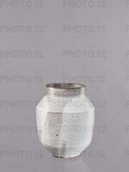 Vase, Nishikawa Minoru b. 1929, ceramicist; vessel, glazed ceramic, Showa period 3rd quarter of the 20th century, Japan, Kyoto; height 19.1 cm, diameter 16 cm., A ceramic vase with a rounded, slightly faceted body and a short neck sits against a plain background; the vase is primarily white with areas of light gray and subtle brownish speckling, the rim and base showing darker gray and brown tones, and the background appears as a muted lavender-gray.