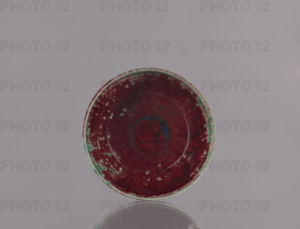 Wan-nari tea bowl wan-nari chawan, Nishizawa Sô ceramicist. Japanese, Showa period 3rd quarter of the 20th century. Ceramic bowl chawan for tea; height 6.3 cm, diameter 14.2 cm., A round shallow ceramic bowl with a predominantly deep red interior and rim that shows mottled patches of green and lighter turquoise, a darker central area with a subtle blue halo and textured red pattern, a glossy reflective surface, and a neutral pale gray background with a faint reflection of the bowl visible below.