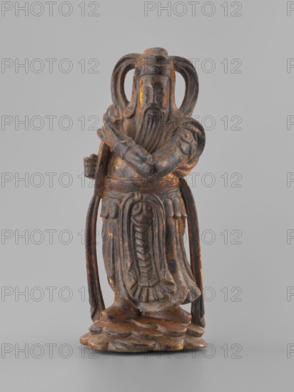 Guardian of Heaven and Defender of the Faith;, figurine; Southeast Asian art; musical instruments; guardians; Vietnamese culture; full wood carving; 18th–19th century; Vietnam Vietnamese–Cambodian border; sculpture; height 33.7 cm, A carved figurine of a robed standing figure with a long beard and a mustache, shown against a plain light gray background. The figure has an elaborate headdress with looped elements extending down on both sides and crossed arms held near the chest. The robe features carved decorative folds and a central vertical motif, and long draping panels fall at the sides. The figure stands on an irregular base that resembles layered rock. The surface shows a worn finish with visible areas of dark brown, black, and spots of gold or ochre highlighting.