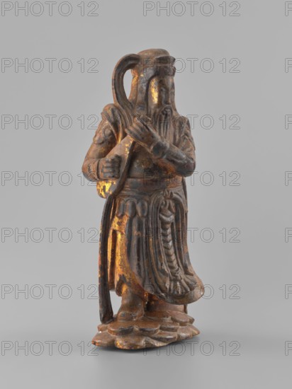 Guardian of Heaven and Defender of the Faith, unknown sculptor, figurine, Southeast Asian art; full wood carving; 18th–19th century; Vietnam Vietnamese–Cambodian border; sculpture; height 33.7 cm; subjects: guardians, musical instruments; culture: Vietnamese., A small carved figure stands on a textured base, rendered in dark brown wood with worn patches of gold and orange highlights; the figure wears layered robes with carved folds and decorative elements, a long beard, and a rounded headdress, and holds a curved object over one shoulder while the other hand rests near the chest, all shown against a plain light gray background.