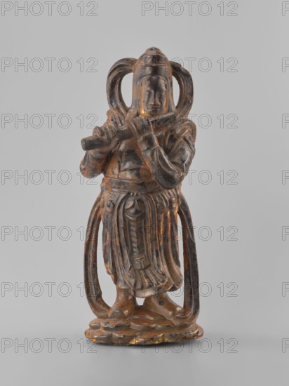 Guardian of Heaven and Defender of the Faith, unknown sculptor; figurine depicting a guardian with a flute; art of Southeast Asia; Vietnamese culture; full wood carving; 18th–19th century; Vietnam Vietnamese-Cambodian border; sculpture; height 33.8 cm, A carved figure standing on a rounded base, depicted holding a horizontal object near its face with both hands; the figure wears a long, flowing garment with carved folds and decorative elements, and has large looped shapes extending from the sides of its head down alongside the body; the surface shows a worn finish with visible patches of brown, dark brown, black, and hints of gold; the background is a plain light gray.