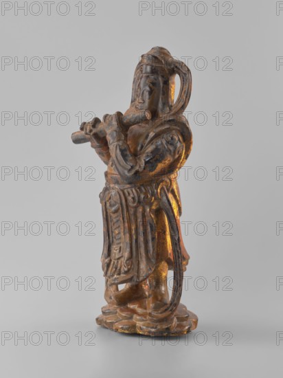 Guardian of Heaven and Defender of the Faith, unknown sculptor; figurine depicting a guardian playing a flute; full wood carving; Vietnamese Vietnamese-Cambodian border origin; 18th–19th century; sculpture; height 33.8 cm., A gilded statue of a robed figure standing on a rounded base, shown in profile, holding a horizontal cylindrical object with both hands near the mouth. The figure wears layered garments with carved folds and ornamental patterns, a sash-like element around the waist, and a looping hair or headdress element extending down the back. The surface shows a worn finish with visible areas of gold and darker brown patina. The background is a smooth neutral gray.