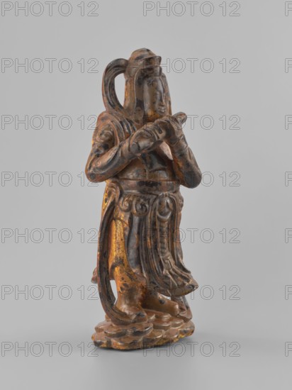 Guardian of Heaven and Defender of the Faith, unknown sculptor; full wood carving; figurine depicting a guardian playing a flute; Vietnamese Southeast Asia culture; 18th–19th century; Vietnam Vietnamese-Cambodian border; sculpture; height 33.8 cm., A carved figurine made of a dark material with worn areas revealing a gold color stands on a textured base; the figure wears flowing robes with carved folds and patterned details, has long hair or a headdress that curves down the back, and holds an object horizontally with both hands near chest level, with the facial features softly modeled and the surface showing patches of dark brown, black, and gold tones.