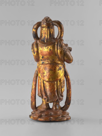 Guardian of Heaven and Defender of the Faith, unknown sculptor; full wood carving figurine; subject: guardian playing a flute; Southeast Asian Vietnamese style; 18th–19th century; origin: Vietnam Vietnamese–Cambodian border; sculpture; height 33.8 cm., A small standing figurine viewed from behind, with a rounded head and a pair of looped elements extending down the sides, wearing a long draped garment that falls to the base and shows folded cloth details; the figure stands on an irregular rounded platform with undulating edges and has a pair of legs visible beneath the hem. The surface shows a mottled, weathered finish with predominant colors of gold, brown, and dark brown or black speckling, and areas where the gold appears worn to reveal darker underlying tones.