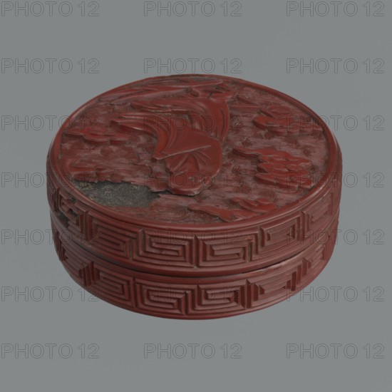 Box with lid, Chinese, Qing dynasty 1644–1911, 19th century. Red lacquer with carved pattern of reeds, meander leiwen, clouds; Buddhist subject matter. Vessel and lid: lid height 2.3 cm, diameter 8.8 cm; vessel height 3.5 cm, diameter 8.9 cm; base height 1.8 cm, diameter 8.8 cm; box height 3.1 cm., A round carved object with a raised decorative lid sits against a gray background; the object is predominantly a deep red with areas of darker red and small patches of black where material is missing, the lid surface features a carved flowing fish-like form surrounded by textured wave-like carvings and floral shapes, and the sides display a repeating geometric stepped pattern in the same red tone.