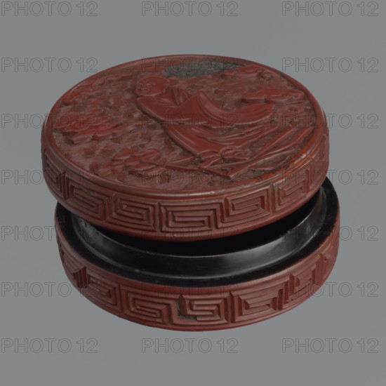 Box with lid, Chinese, Qing dynasty 1644–1911, 19th century. Red lacquer, carved pattern of reeds, meander leiwen, clouds; Buddhist subject matter. Vessel and lid: lid height 2.3 cm, diameter 8.8 cm a; vessel height 3.5 cm, diameter 8.9 cm; base height 1.8 cm, diameter 8.8 cm b; box height 3.1 cm., A round carved container with a textured circular lid featuring a raised figure in flowing robes and decorative swirling patterns, the top surface and sides showing carved geometric border motifs; the visible colors are red, black, and a few darker brownish areas, set against a neutral gray background.