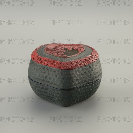 Box with a lid, Chinese, Qing 1644–1911, 19th century. Two-tone lacquer with carved decoration depicting figurative scenes: peaches, flowers, bats, pines, rocks, clouds, kikko pattern. Container, height 7.3 cm, width 10.6 cm, depth 10.5 cm., A rounded, textured container with a muted green surface and a decorative red band around its top edge sits on a neutral light-gray background; the container has a patterned, slightly rough exterior and a lid topped with an ornate red raised design, showing a contrast between the green body and the red decorative elements.