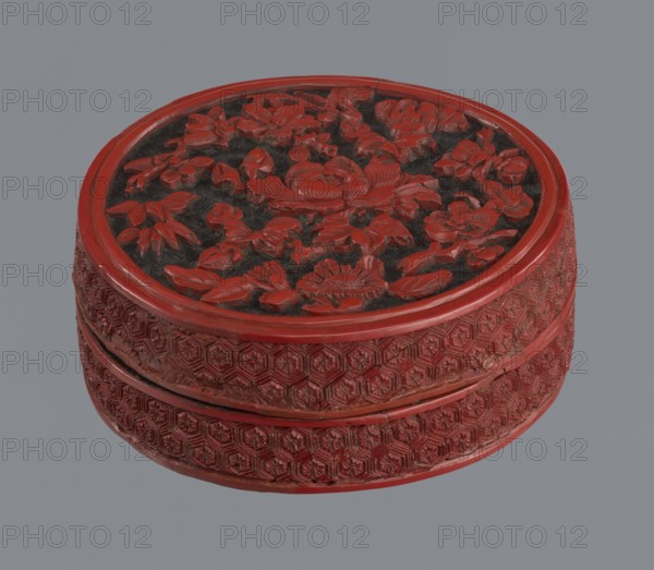 Box with lid, maker, Chinese; Qing dynasty 1644–1911, 19th century. Two-tone lacquer, red and black black lacquer inside. Round container decorated with floral ornament—peonies, bamboos, peaches—and kikko geometric pattern. Dimensions: base height 2.1 cm, diameter 8.8 cm a; lid height 1.8 cm, diameter 8.8 cm b; box height 3.8 cm, diameter 8.9 cm., A round carved object with a raised floral relief on its top and a patterned band around its sides, primarily red with dark recessed areas in the top relief; the top shows intricately carved flowers and leaves in red against a dark background, and the sides display repeating geometric floral motifs in red; the background behind the object is a flat gray.