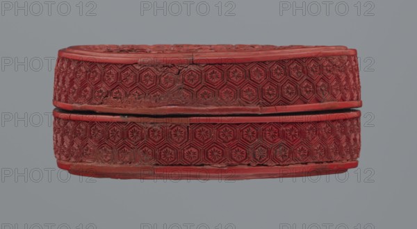 Box with lid, Chinese, Qing 1644–1911, 19th century. Two-tone lacquer, red and black black lacquer inside. Decoration: floral ornament with peonies, bamboos, peaches iconography, kikko geometric pattern. Container; base: height 2.1 cm, diameter 8.8 cm; lid: height 1.8 cm, diameter 8.8 cm; box height 3.8 cm, diameter 8.9 cm., A carved circular object with an oval profile sits against a gray background; the object is red with areas of darker red and some spots of brownish wear, and it has a repeating geometric floral pattern carved around its sides and top, with a narrow horizontal separation around its middle showing a thin dark gap.